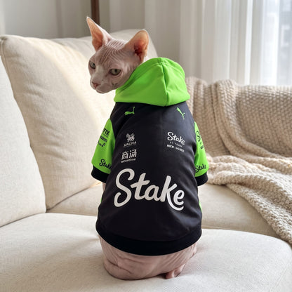 Sphynx cat wearing black green Stake F1 Sauber pet hoodie on sofa with white Stake logo on back